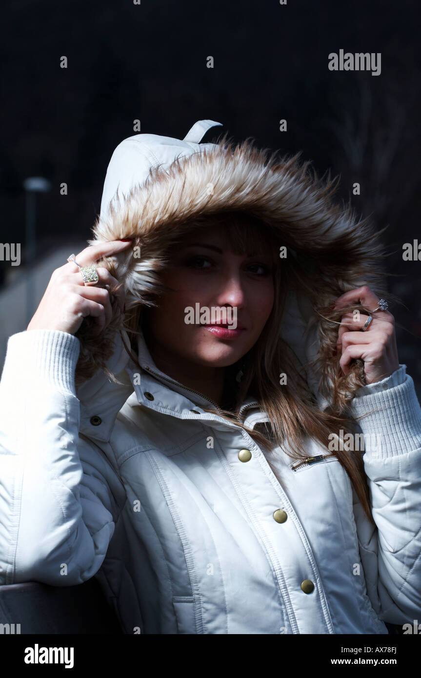 Blonde Frau, die eine weiße Jacke mit einer Kapuze mit Pelzbesatz trägt, die Kapuze mit ihren Händen justiert, nachts draußen. Stockfoto