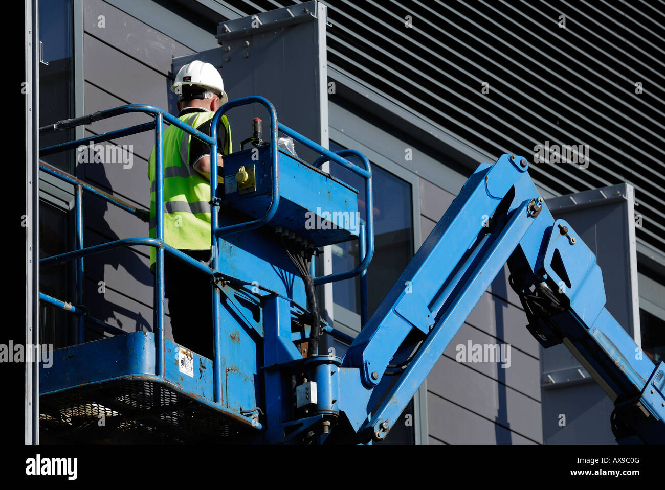 Ein Bauarbeiter in einem Aufzug, der einen Schutzhelm und eine Sicherheitsweste trägt, arbeitet an der Außenseite eines Gebäudes mit hydraulischem Aufzug oder Kirschpflücker Stockfoto