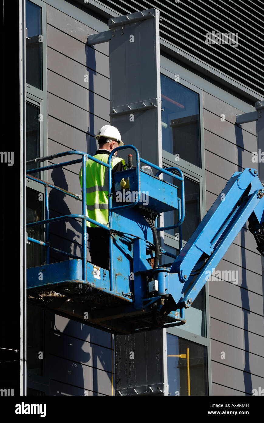 Ein Bauarbeiter in einem Aufzug, der einen Schutzhelm und eine Sicherheitsweste trägt, arbeitet an der Außenseite eines Gebäudes mit hydraulischem Aufzug oder Kirschpflücker Stockfoto