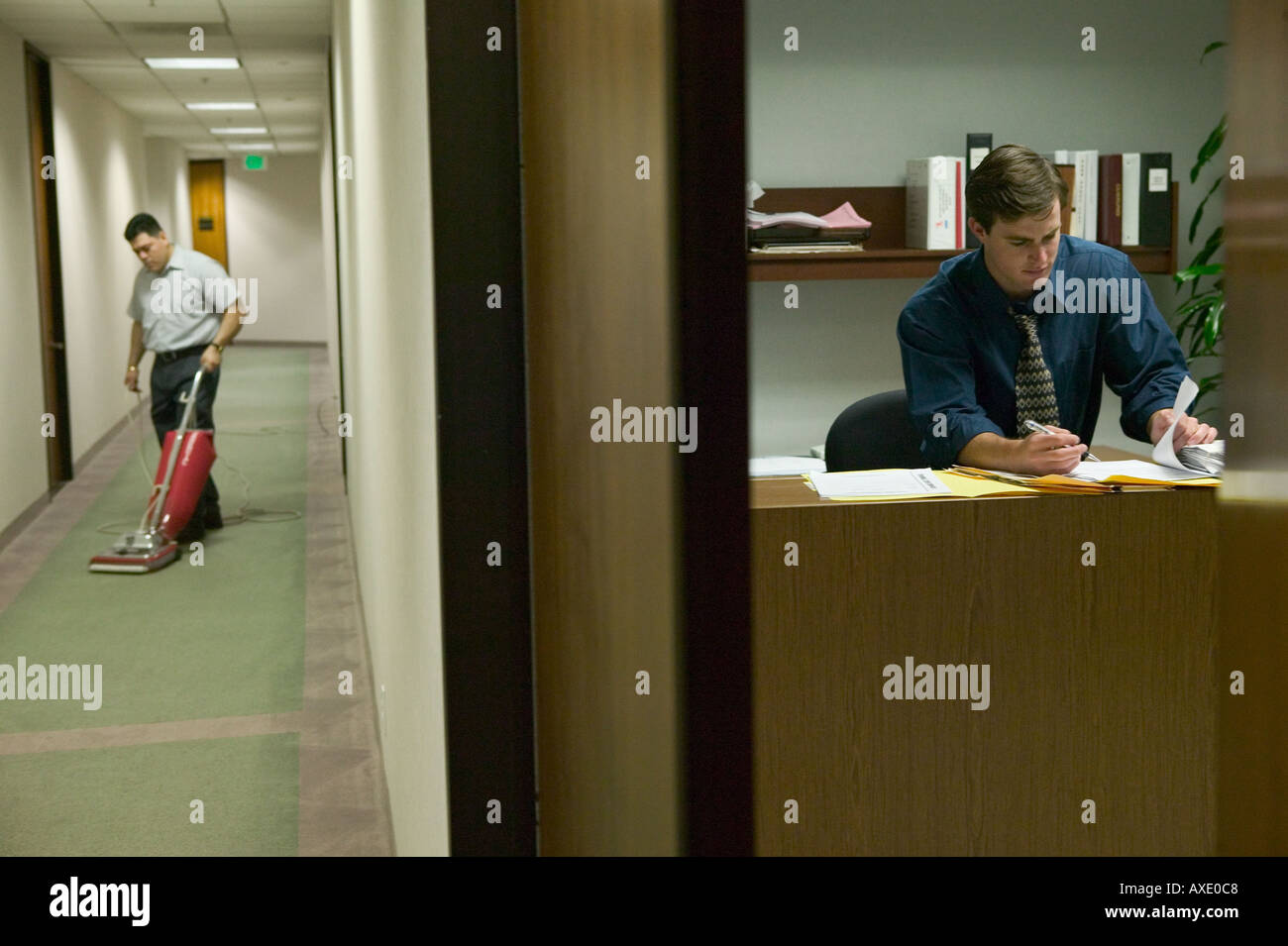 Arbeitnehmer arbeitet nach Stunden im Büro während der Hausmeister Vacuumes Büro Flur Stockfoto