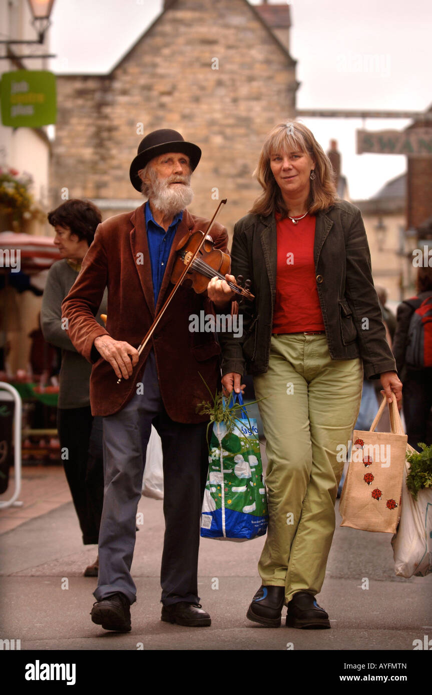 EINE ELDERLEY MUSIKER SPIELT EINE GEIGE, DIE BEGLEITUNG EINER DAME EINKAUFEN AUF DEM BAUERNMARKT IN STROUD GLOUCESTERSHIRE UK Stockfoto