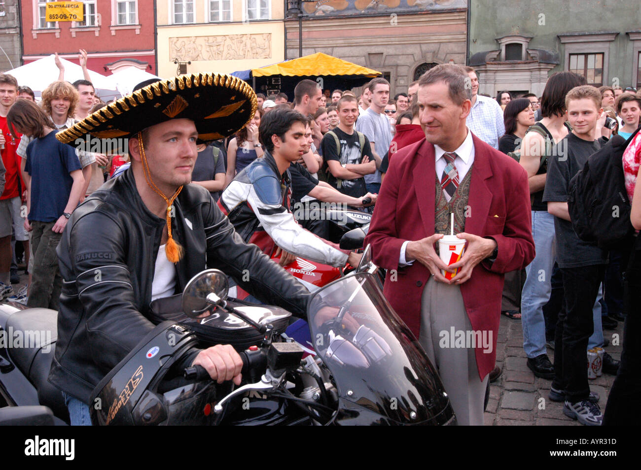 Junge Rockfans treffen sich auf einem Stadtplatz, ein Mann in einem Sombrero sitzt auf einem Motorrad, während andere, darunter ein Mann in einem roten Blazer, zuschauen. Stockfoto