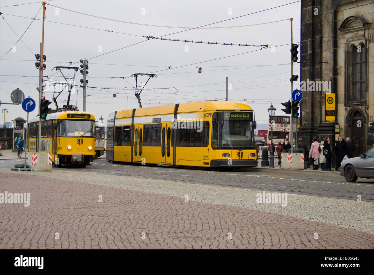 Zwei gelbe Straßenbahnen an einer Haltestelle in Dresden, Deutschland, mit Kopfsteinpflasterstraßen, wartenden Passagieren und sichtbaren Oberbahnkabeln. Stockfoto