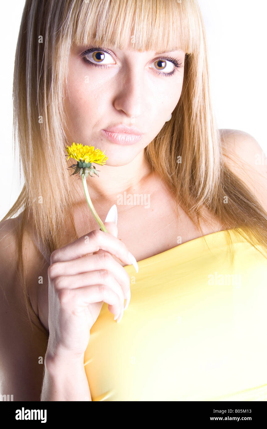 Blonde Frau, die eine gelbe Blume nahe ihrem Gesicht hält, ein gelbes Kleid trägt, fotografiert in einem Atelier mit weißem Hintergrund. Stockfoto