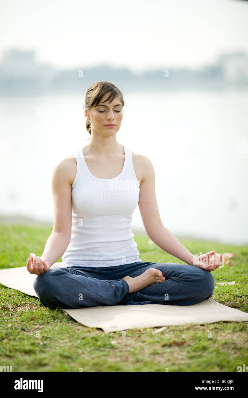 Eine junge Frau sitzt im Lotussitz auf eine Yoga-Matte in einem Park. Stockfoto