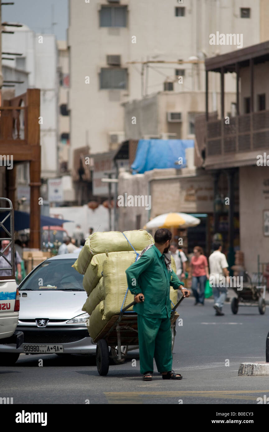 Dubai, Vereinigte Arabische Emirate (VAE). Ein Portier ziehen einem schwer beladenen Karren durch viel Verkehr entlang der Uferpromenade in der Altstadt Stockfoto