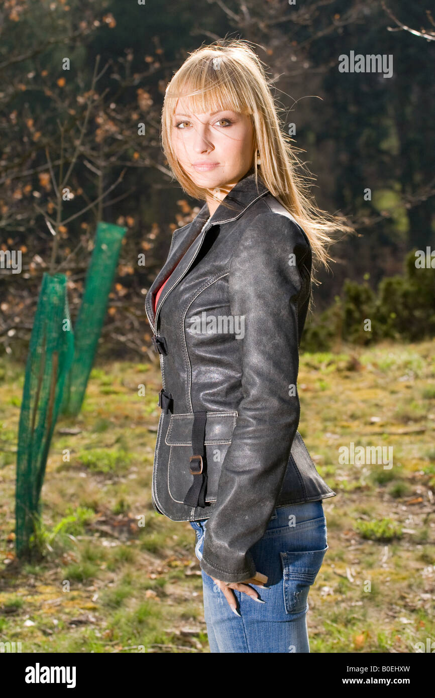 Blonde Frau in Lederjacke und Jeans, die draußen steht und mit Wind geblasenen Haaren über die Schulter blickt, in einer natürlichen Waldlandschaft. Stockfoto