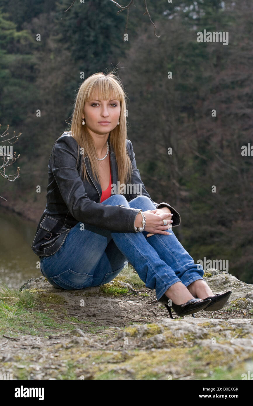Blonde Frau in Lederjacke und Jeans, die im Kreuz auf einer felsigen Oberfläche in der Nähe eines Waldflusses sitzt und direkt in die Kamera blickt. Stockfoto