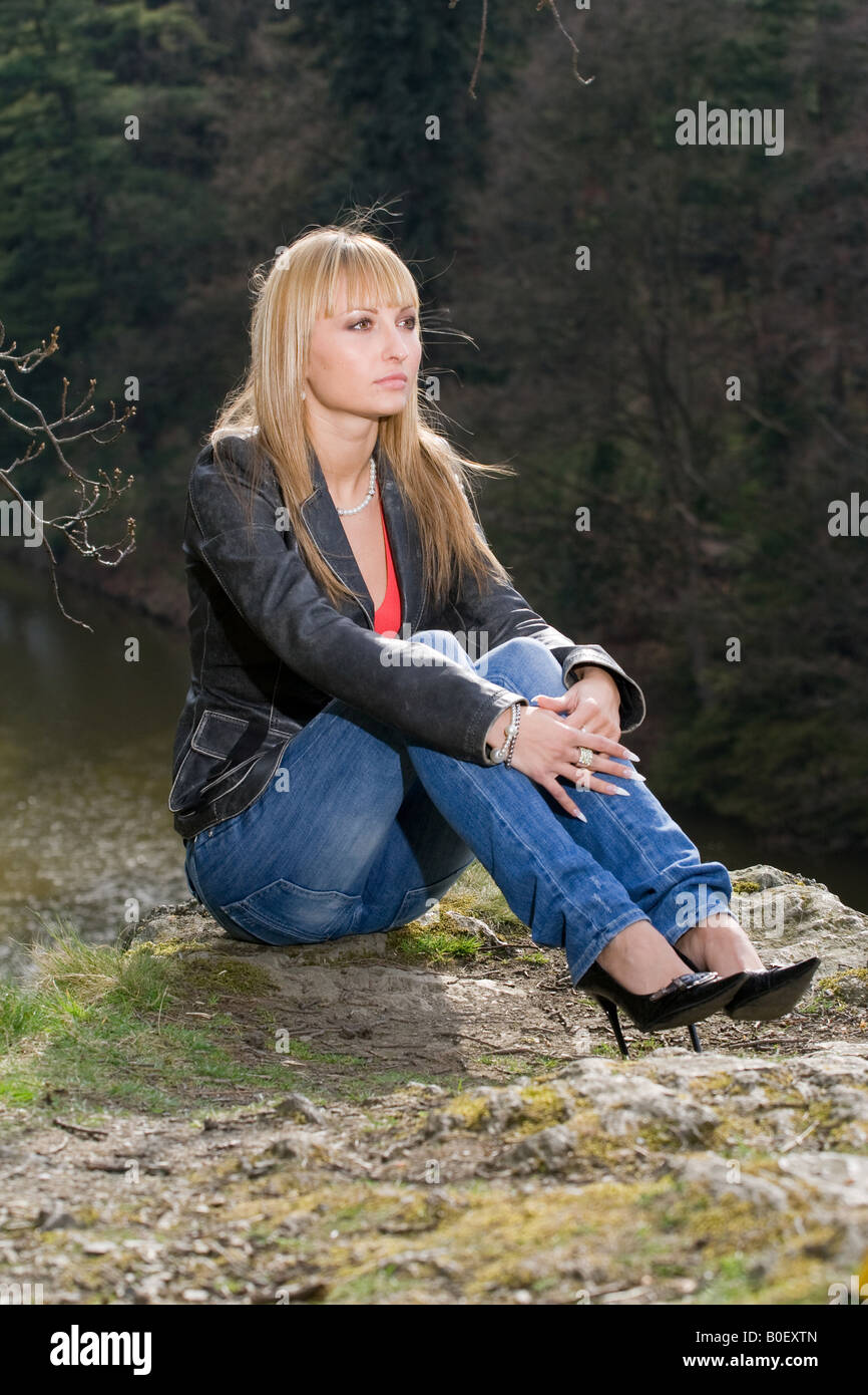 Blonde Frau in einer Lederjacke, die auf einem Felsen in der Nähe eines ruhigen Flusses sitzt und nachdenklich in die Ferne blickt, in einer natürlichen Umgebung im Freien. Stockfoto