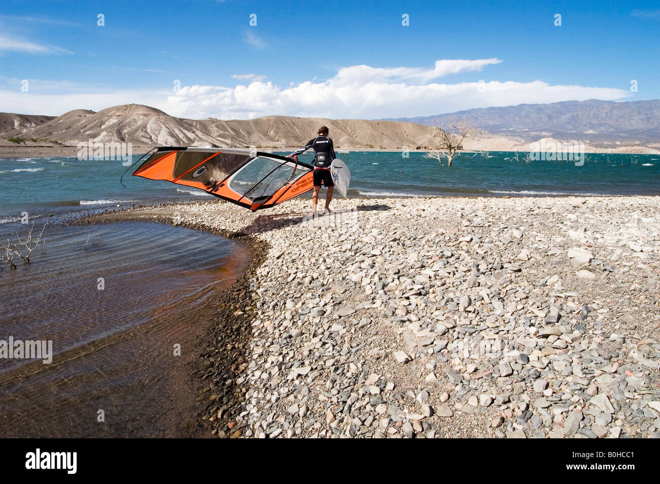 Windsurfer im Neoprenanzug mit seinem Board und Segel über den trockenen Kies windgepeitschten Seeufer umgeben von Bergen, Cuesta d Stockfoto