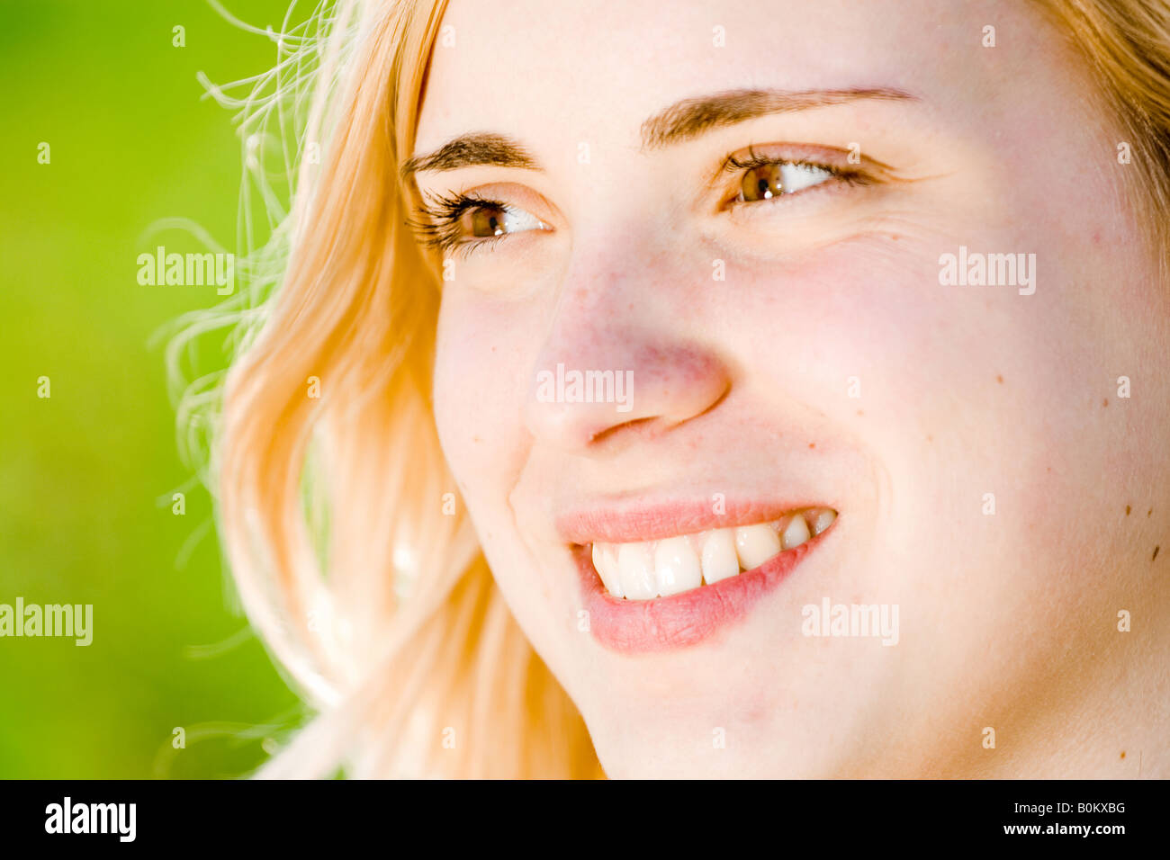 Nahaufnahme einer lächelnden blonden Frau mit heller Haut und Sommersprossen, die in sanftem Sonnenlicht mit grünem, verschwommenem Hintergrund wegschaut. Stockfoto