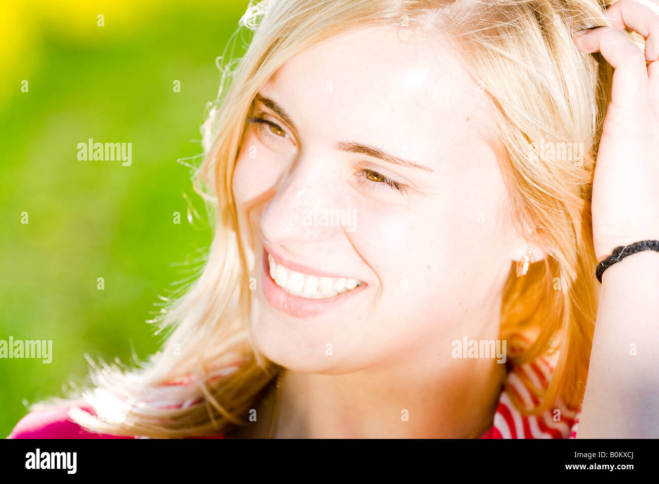 Lächelnde blonde Frau mit heller Haut, trägt ein rosa gestreiftes Hemd, berührt ihr Haar, draußen im Sonnenlicht mit einem leuchtend grünen Hintergrund. Stockfoto