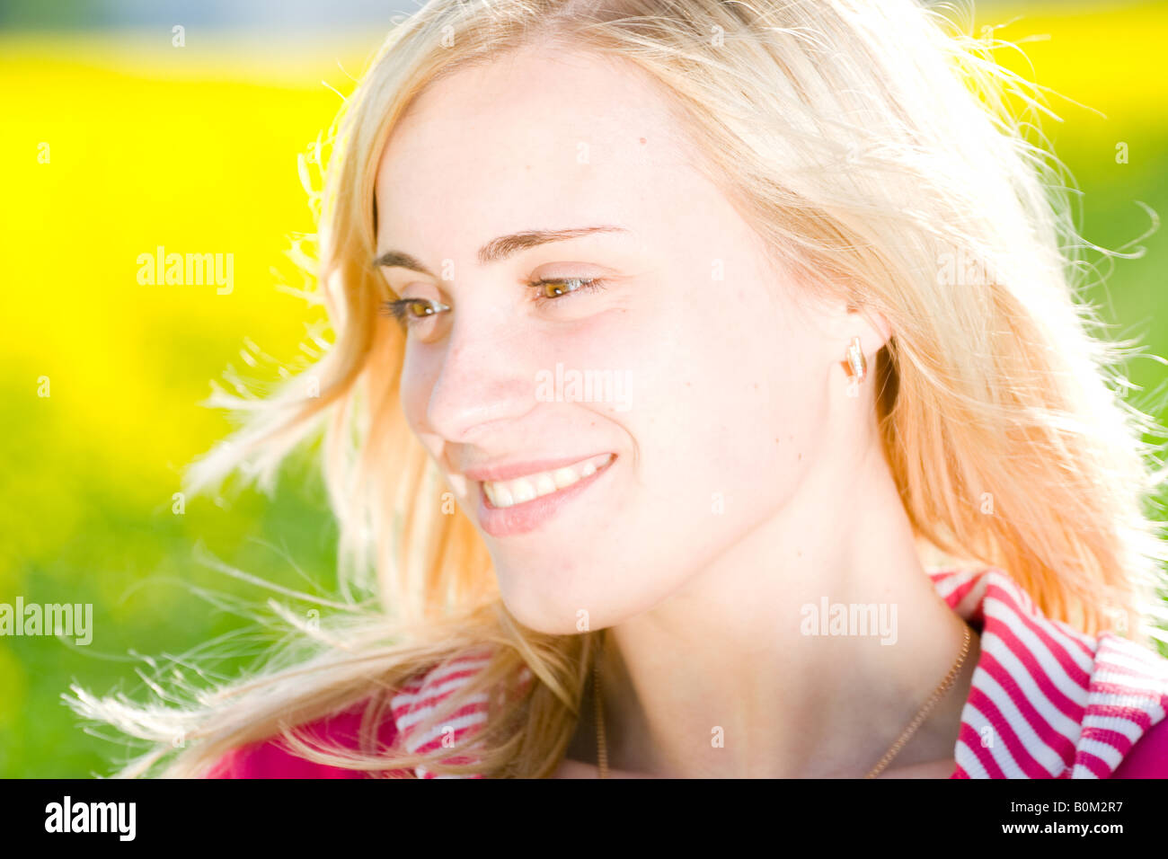 Lächelnde blonde Frau mit heller Haut, trägt ein rosa gestreiftes Hemd, berührt ihr Haar, draußen im Sonnenlicht mit einem leuchtend grünen Hintergrund. Stockfoto