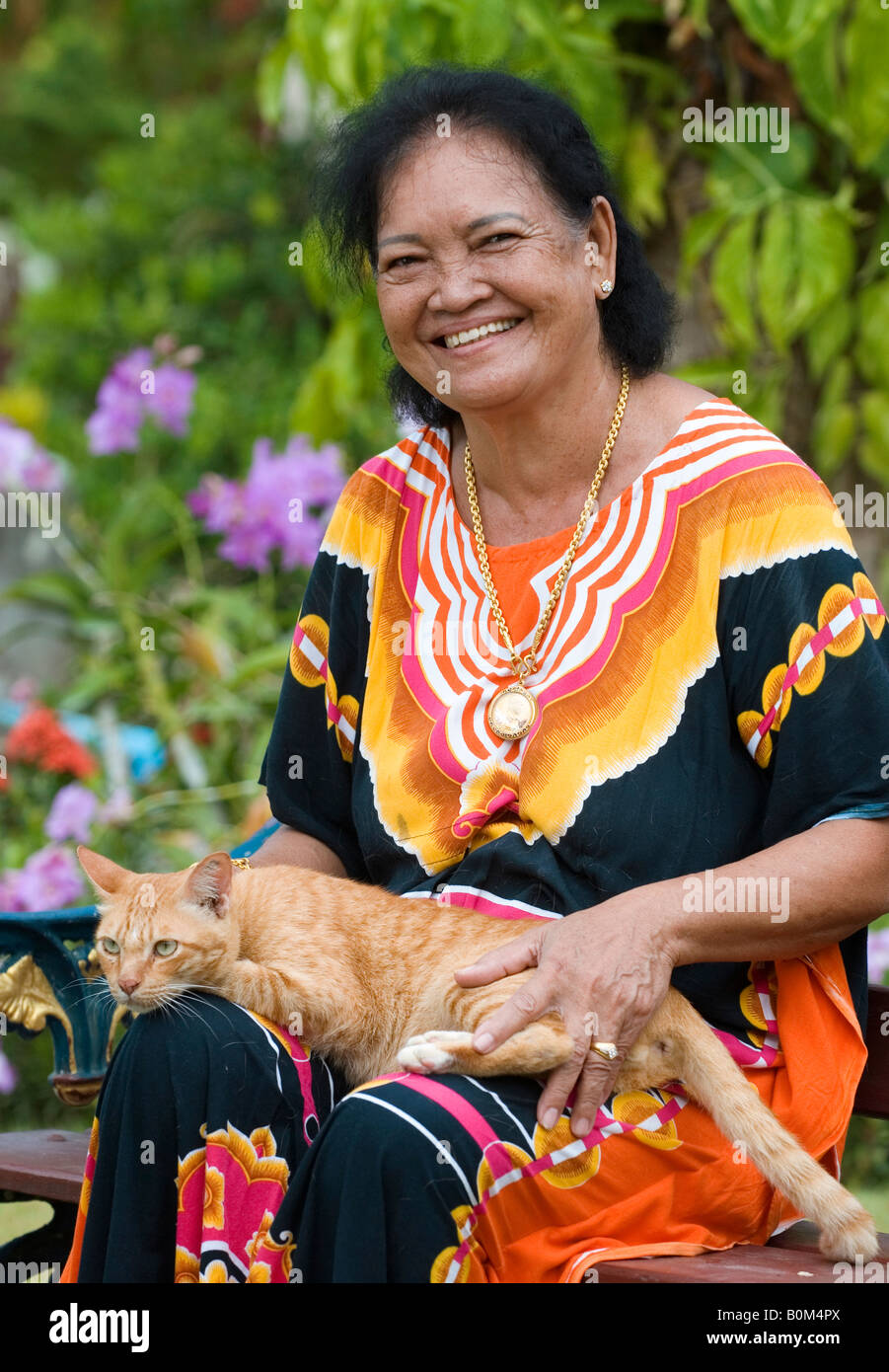Eine bunt gekleideter älterer Thai Dorf Frau genießt sitzen in ihrem Garten mit Tabby Katze, Süd-Thailand. Stockfoto