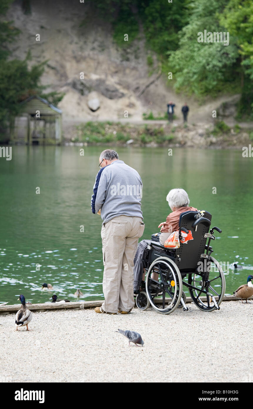 Ein älterer Mann mit Frau im Rollstuhl, der Enten am Swanbourne Lake, Arundel, West Sussex, England, Großbritannien füttert Stockfoto