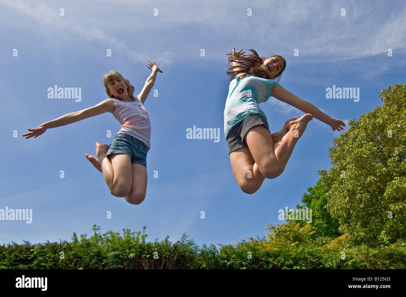 Zwei junge Mädchen (10 und 13 Jahre) springen von einem Trampolin in der Luft an einem sonnigen Tag. Stockfoto