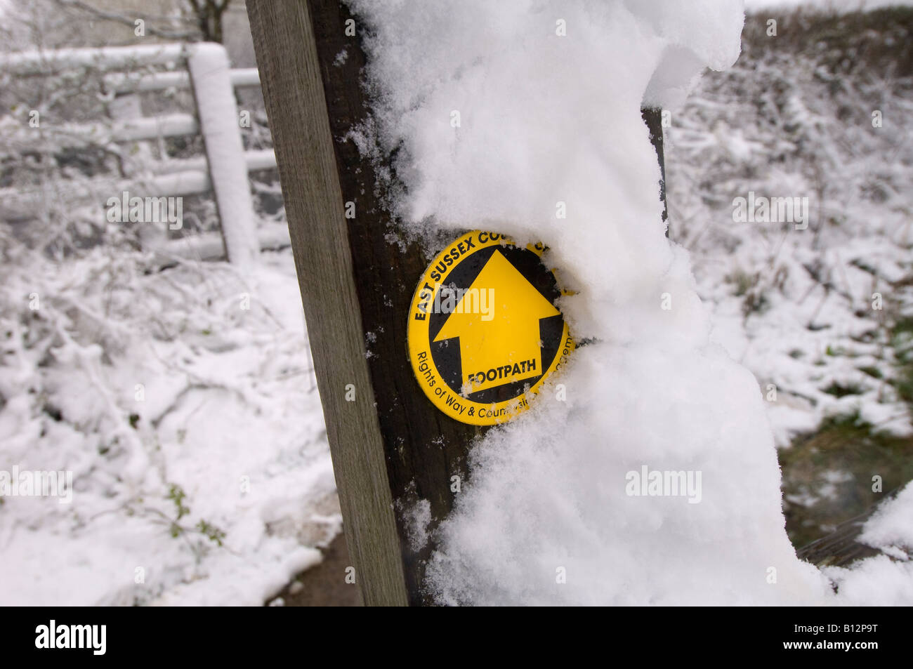 Winterspaziergang: Ein schneebedeckter Pfosten und ein Eisenbahnzaun mit gelbem Wegweiser in Sussex. Stockfoto