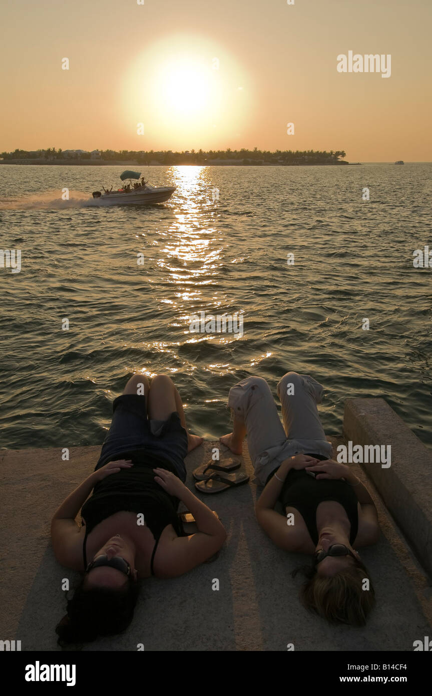Zwei Freundinnen zurücklehnen am Mallory Square Pier als Menschen Welle von vorbeifahrenden Boot bei Sonnenuntergang, Key West, Florida Stockfoto