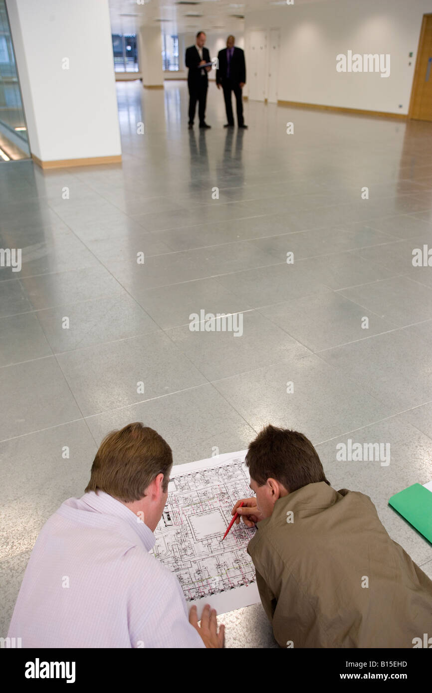 Zwei Ingenieure überprüfen einige Pläne, wie zwei Männer in Anzügen eine Zwischenablage im Hintergrund lesen. Stockfoto