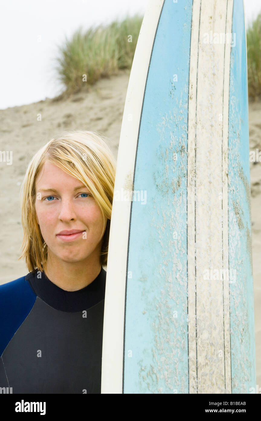 Blonde surfer Frau in Neoprenanzug mit blauem Surfbrett mit einem pazifischen Nordwesten Strand im Hintergrund. Stockfoto