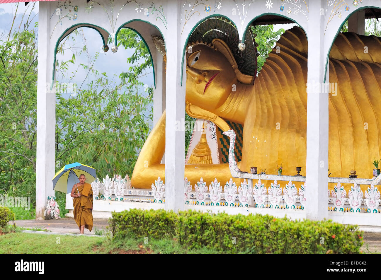 Ein senior buddhistischer Mönch mit Regenschirm vor liegenden Buddha. Sangkhla Buri, Thailand. Stockfoto