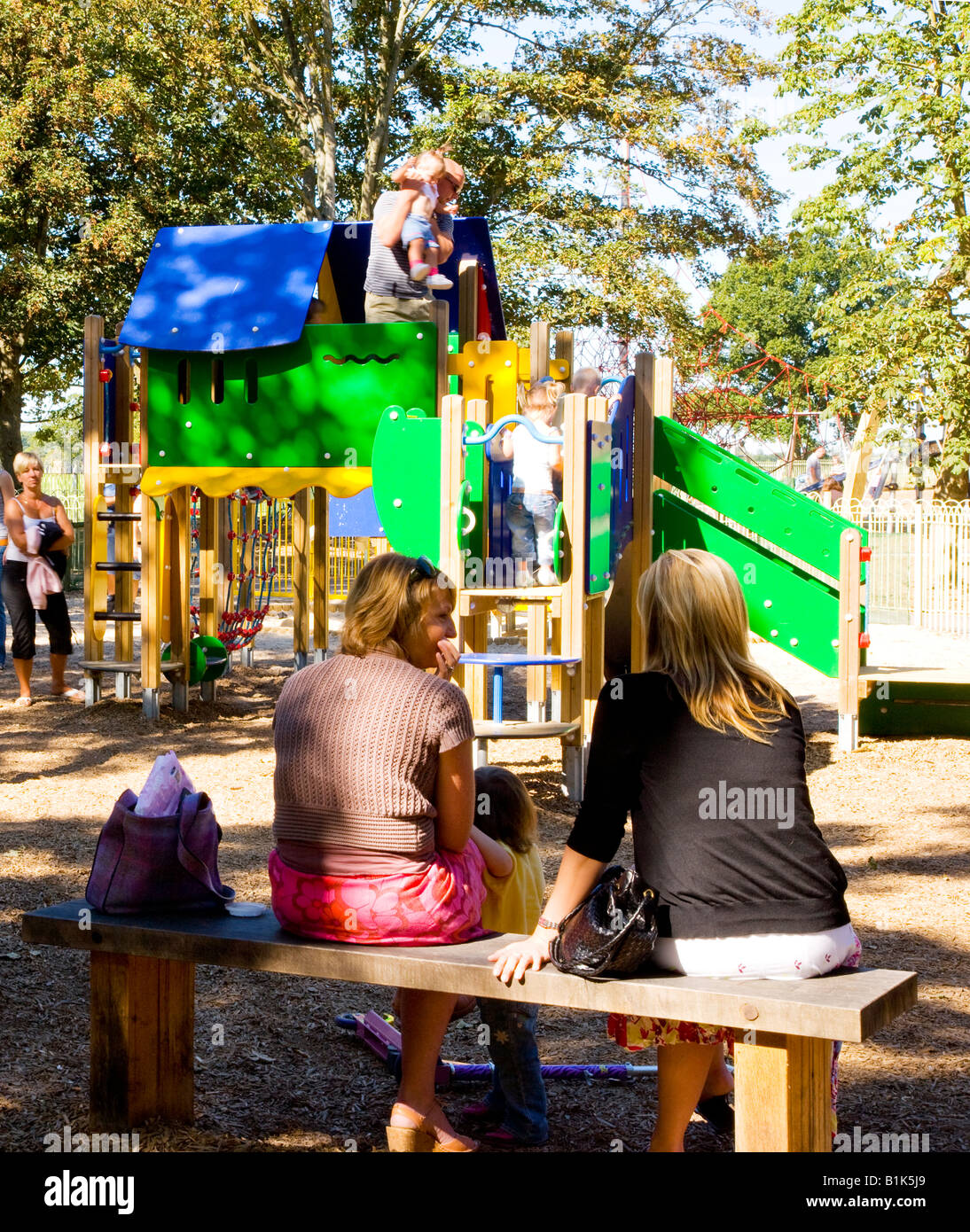 Zwei Mütter, die im Chat, wie sie ihre Kleinkinder auf helle, moderne Abenteuer Spielgeräte im Lydiard Park, Swindon spielen beobachten Stockfoto