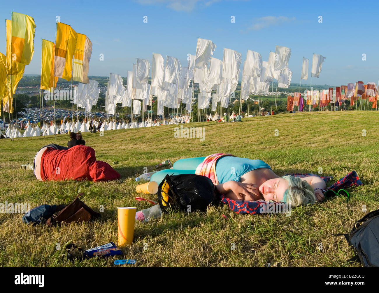 Zwei junge Festivalbesucher schlafen auf dem Rasen mit Blick auf dem Glastonbury Festival-Gelände in der Ferne. Stockfoto