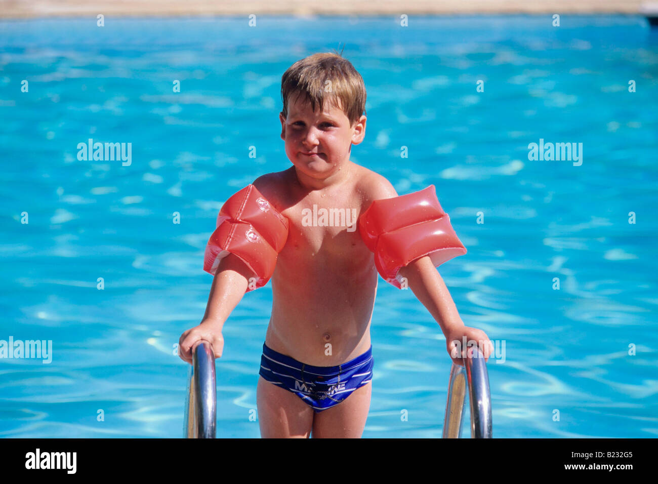 Ein kleiner Junge steht auf den Stufen des einen Pool in der Sonne direkt in die Kamera tragen Arm Flotation aids Stockfoto