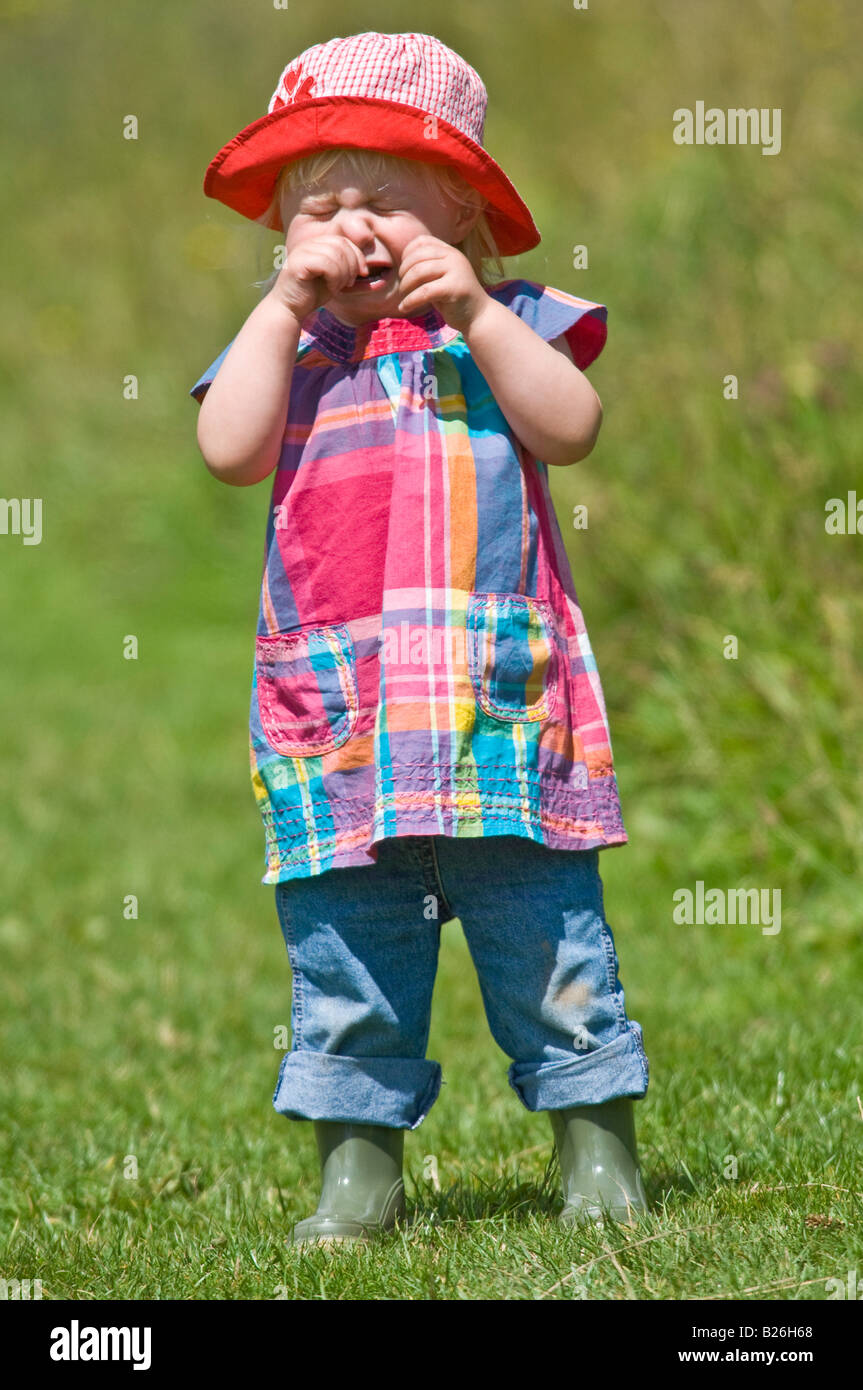 Eine süße junge Mädchen Kleinkind weint tragen Gummistiefeln und rotem Sonnenhut an einem sonnigen Tag in einem grünen Feld. Stockfoto