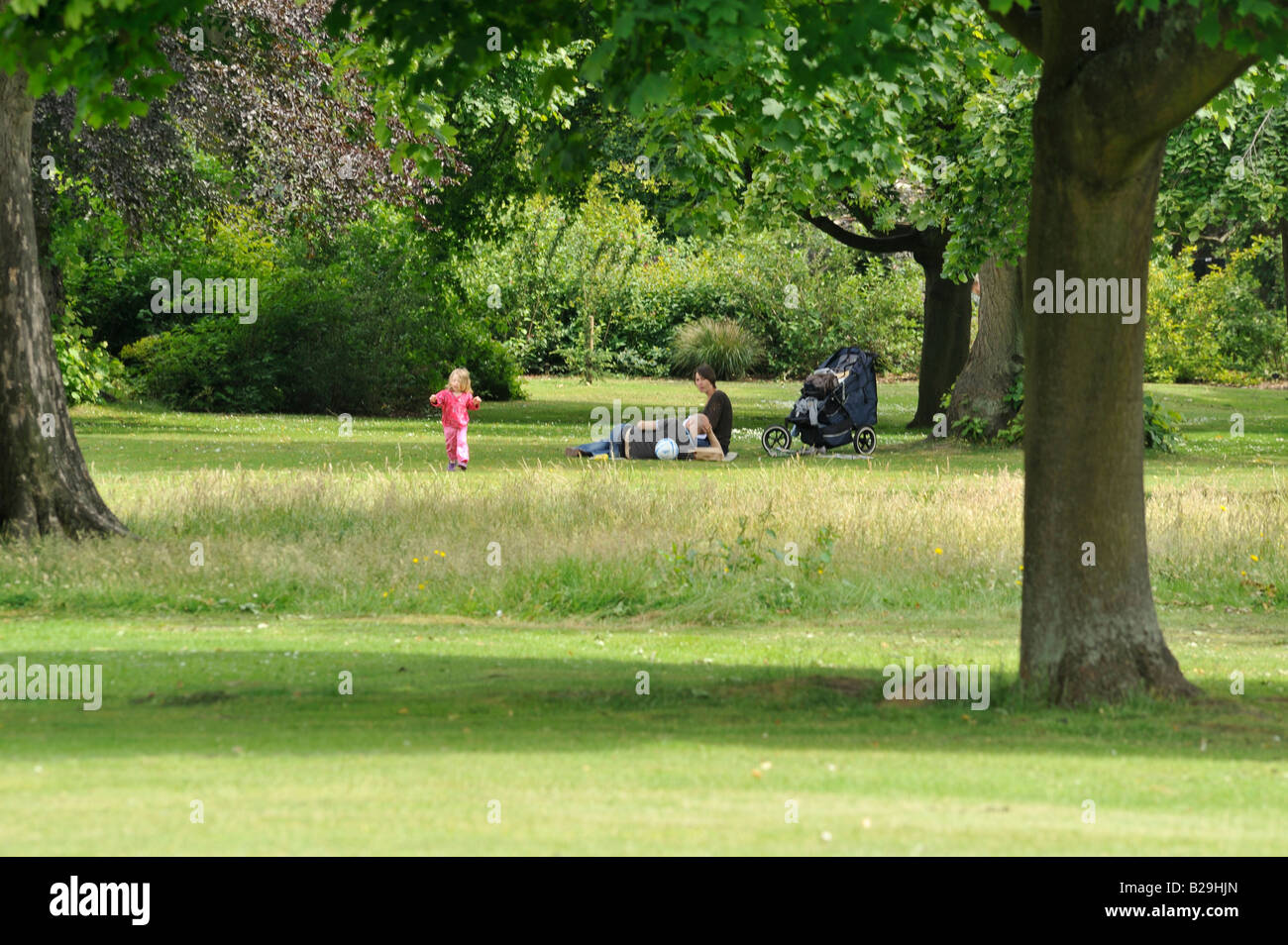 Menschen im Park: Mama und Papa entspannen, während ihr Kind glücklich auf den grasbewachsenen Freiflächen spielt. Stockfoto