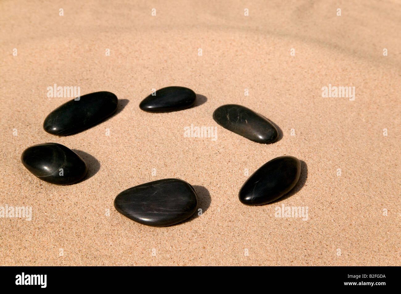 Sechs schwarze glatte Kieselsteine in einem Kreis auf einem sandigen Strand angeordnet Stockfoto