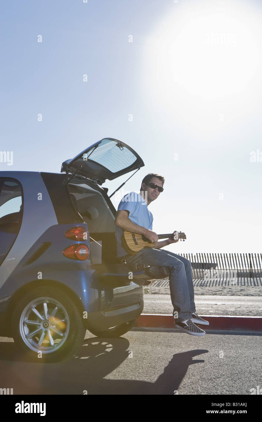 Junger Mann sitzt beim Start des Smartcar am Strand, die Gitarre zu spielen Stockfoto