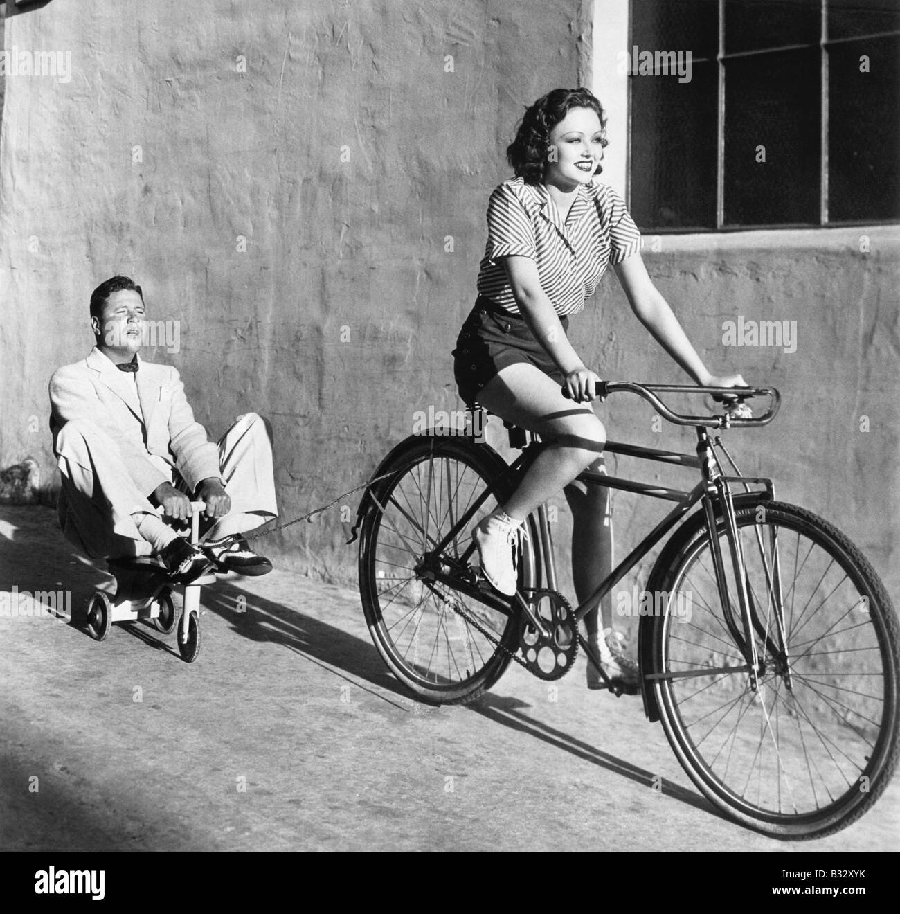 Frau auf einem Fahrrad ziehen einen erwachsenen Mann auf einem Spielzeug Dreirad Stockfoto