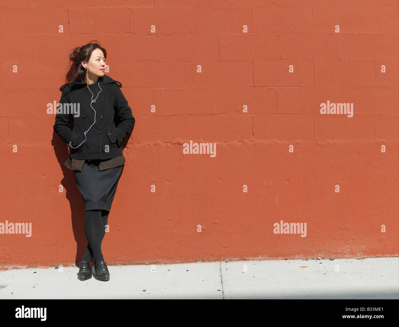 Eine junge Frau auf ihrem Handy hört und schaut Weg stehend gegen eine Wand aus rotem Backstein im winter Stockfoto