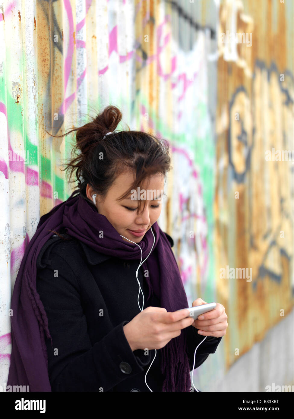 Eine junge Frau trägt einen Mantel hört Musik auf ihrem Mobiltelefon stehend gegen eine Wand graffitied Stockfoto
