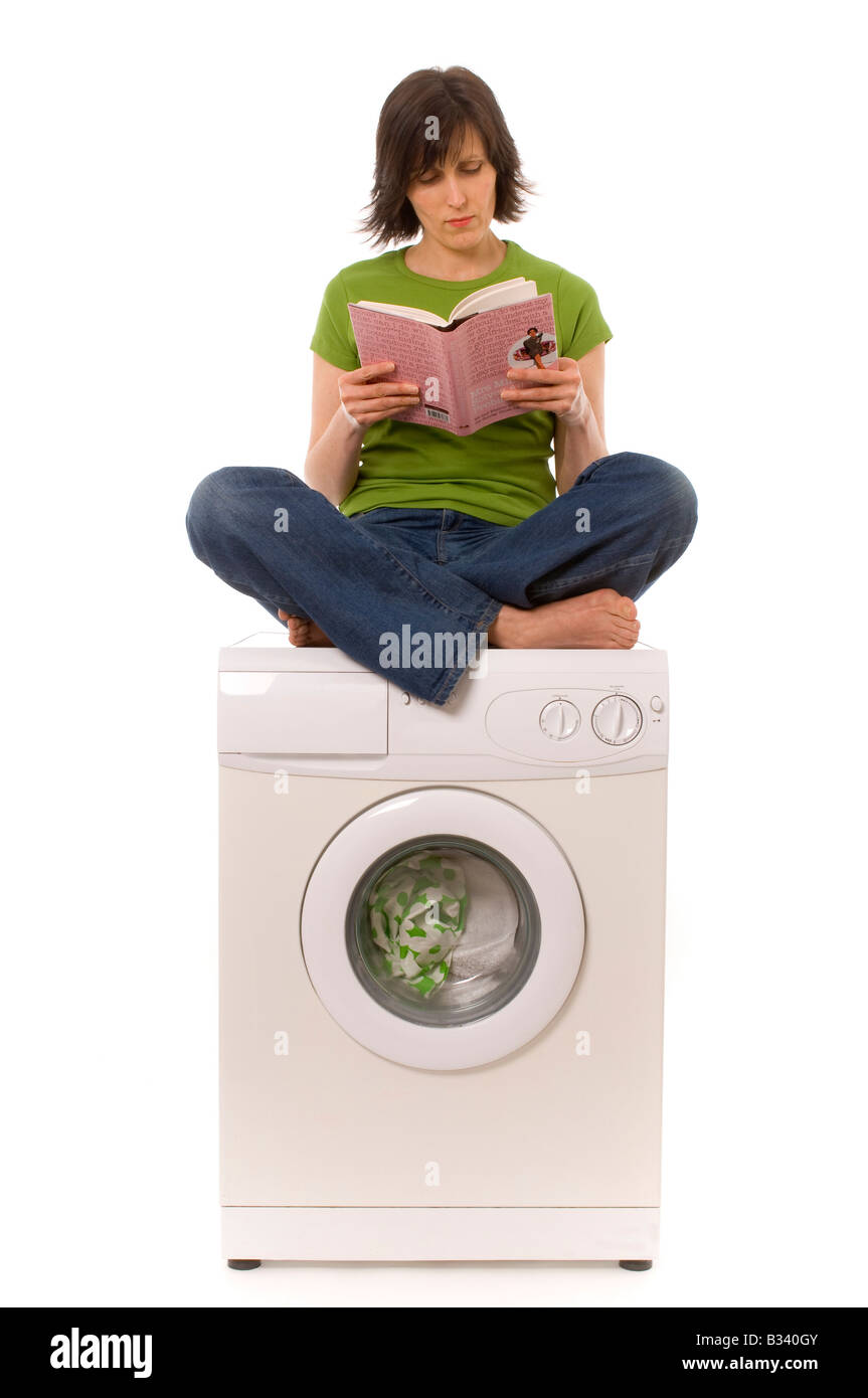 Weiße Frau mit dunklen Haaren, mit grünem Oberteil und blauer Jeans. Sie sitzt kreuzbeinige Beine auf einer Haushaltswaschmaschine und liest ein Buch. Stockfoto