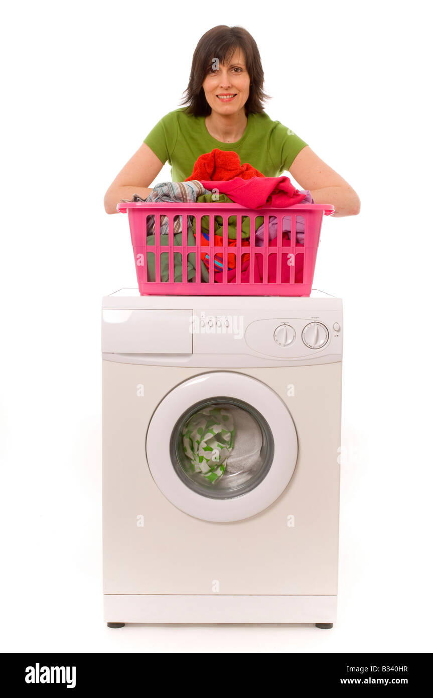Lächelnde junge weiße Frau, die ein grünes T-Shirt trägt und sich auf einen pinkfarbenen Wäschekorb voller Kleidung lehnt, der sich auf einer Waschmaschine befindet Stockfoto