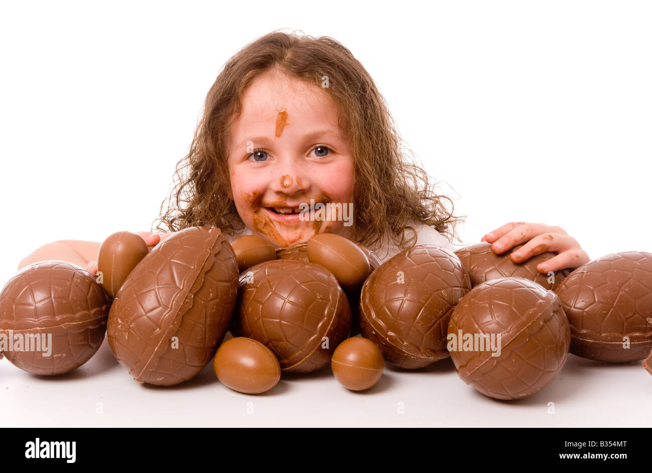 Ein kleines weißes Kind mit geschmolzener Schokolade im Gesicht und an den Händen, das hinter einem großen Haufen Ostereier auftaucht, vor weißem Hintergrund. Stockfoto