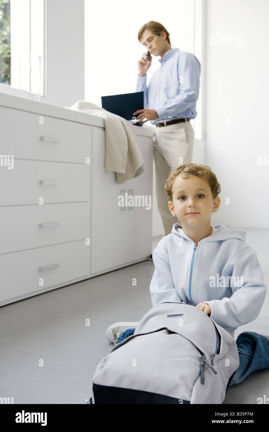 Kleiner Junge sitzt auf dem Boden mit Rucksack, Vater mit Handy im Hintergrund Stockfoto