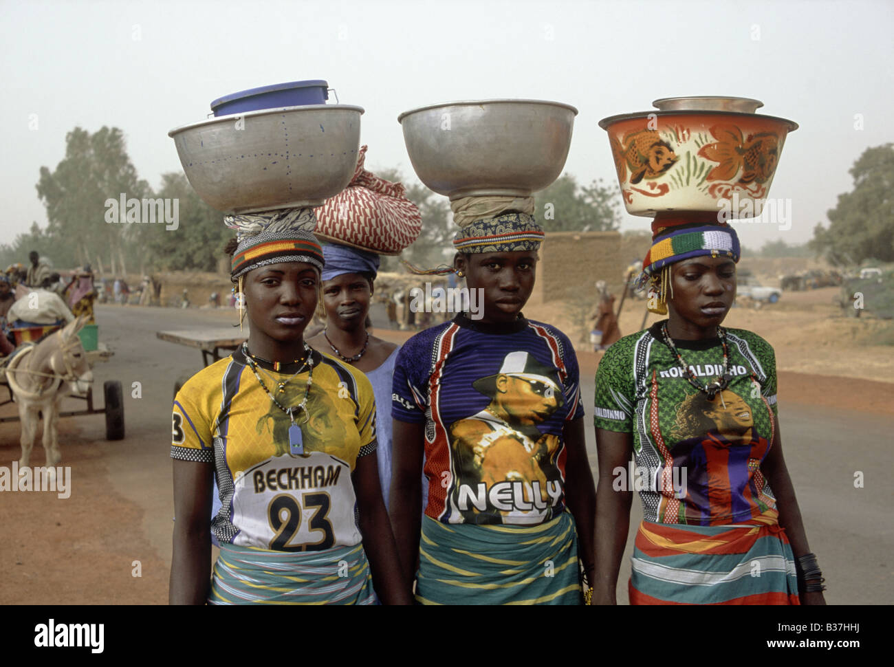 Drei junge Frauen mit westlichen Stil Teeshirts Namen Beckham Nelly Ronaldinho Lasten auf Kopf SOMADOUGOU MALI Sonntagsmarkt Stockfoto