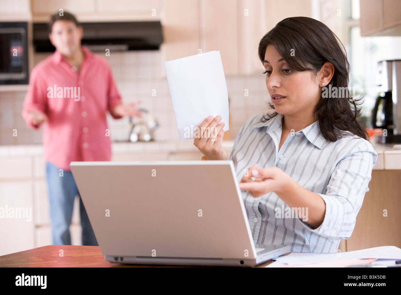Frau in der Küche mit Papierkram mit Laptop mit Mann im Hintergrund Stockfoto