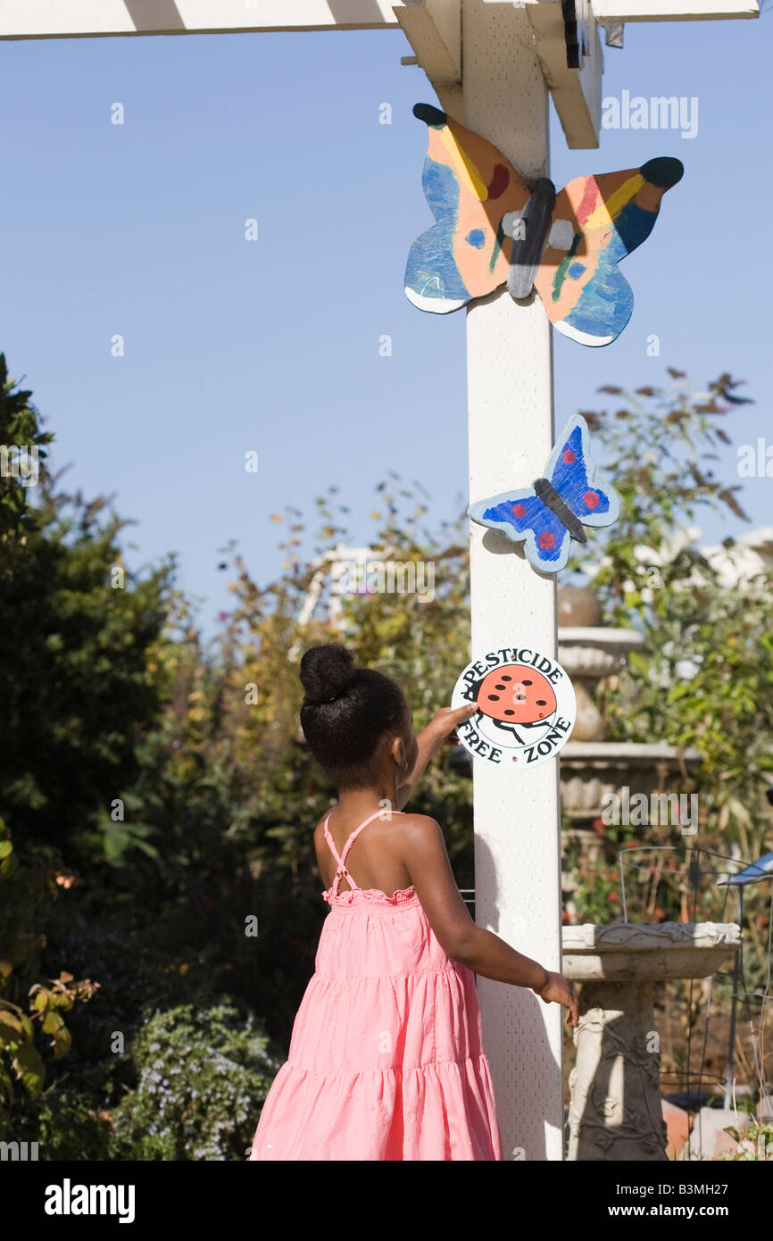 Eine Mischrasse kleines Mädchen inspiziert das "Pestizid freie" Marienkäfer Schild an einer organischen einheimische Pflanze Garten. Stockfoto