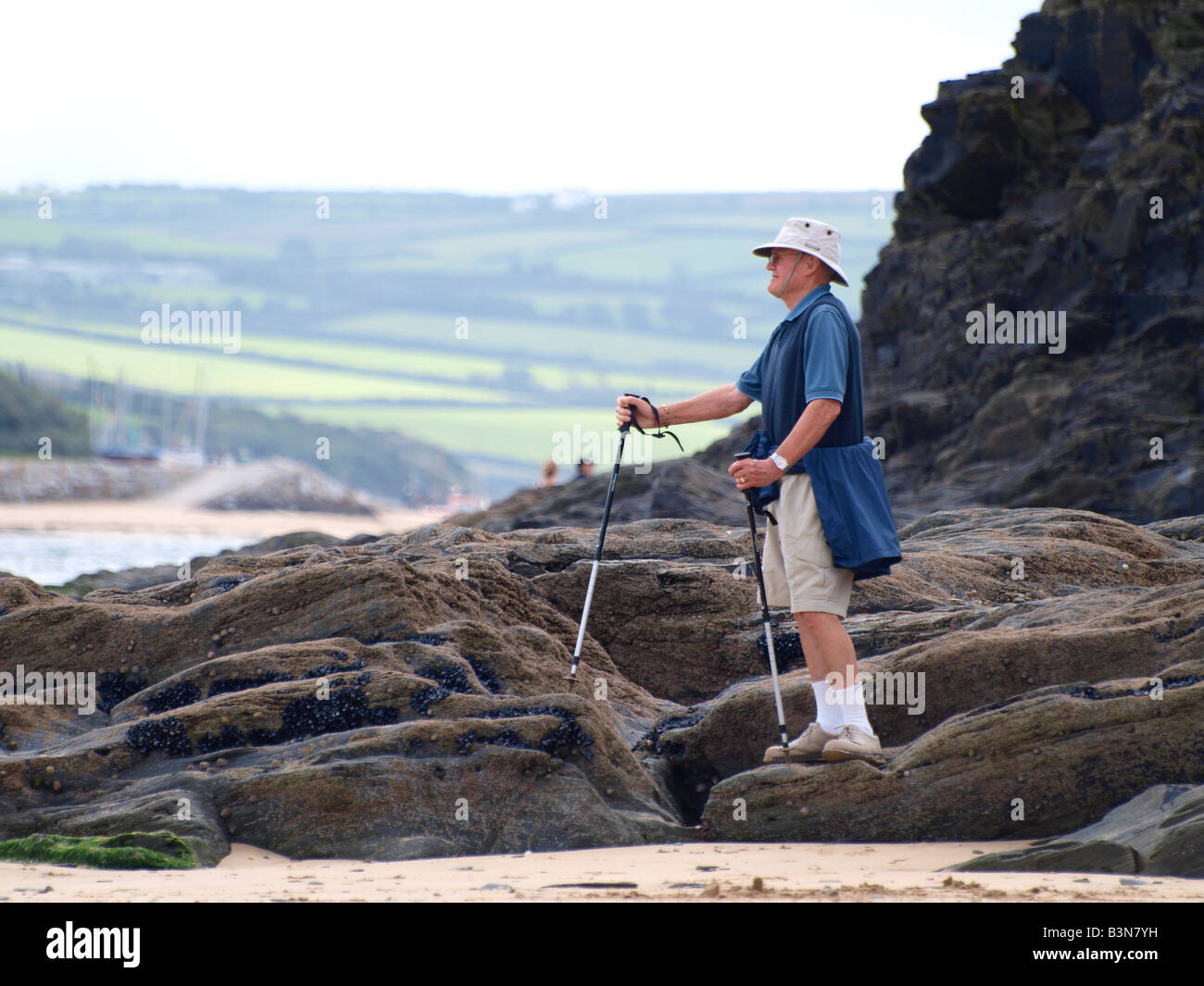 50er Jahre plus Mann auf Felsen mit Blick auf das Meer mit Wanderstöcken für Unterstützung. Stockfoto