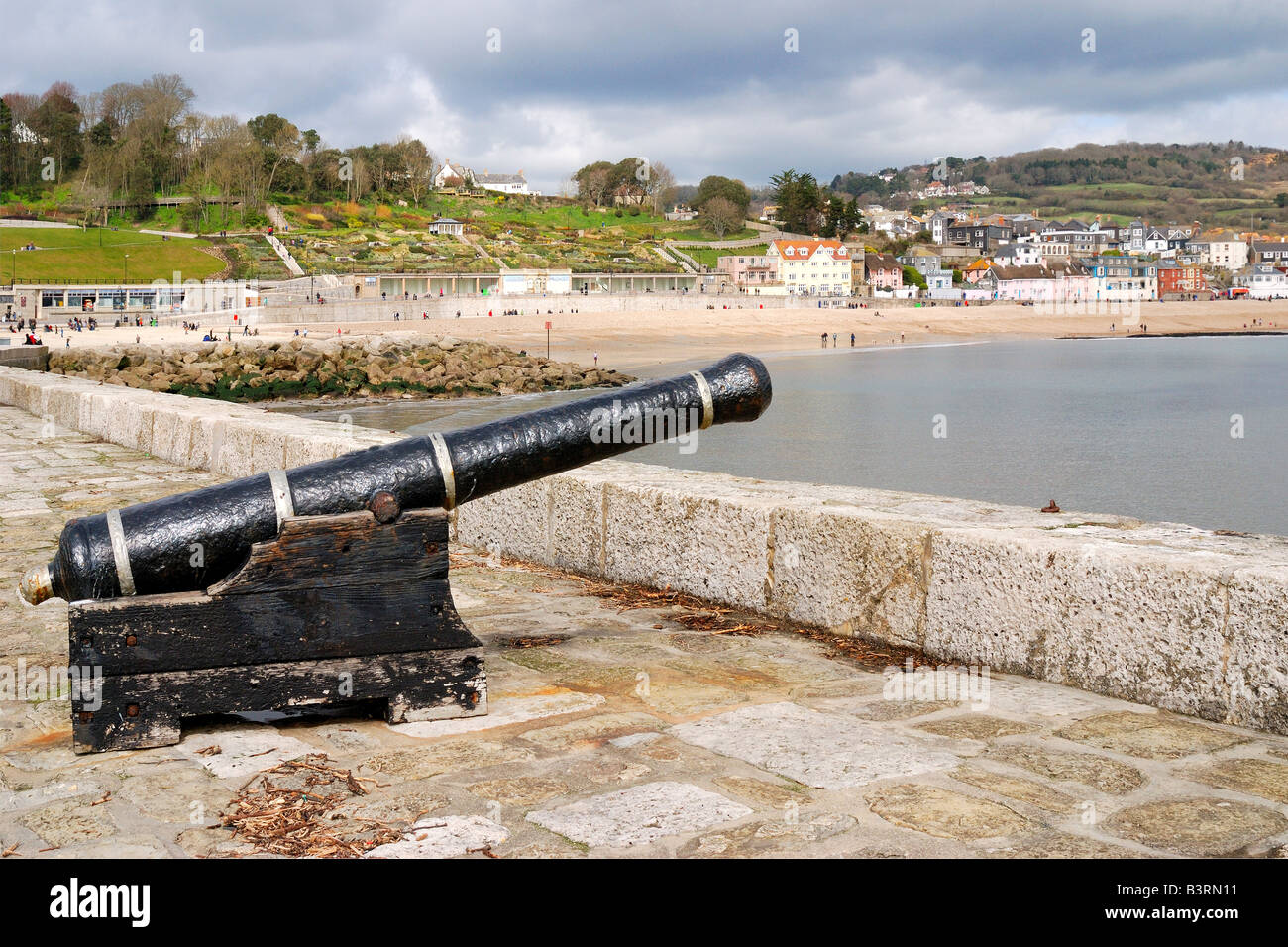 Gedenk-Kanone auf der Hafenmauer in Lyme Regis in Dorset mit strahlendem Sonnenschein und einem dunklen bewölkten Himmel Stockfoto