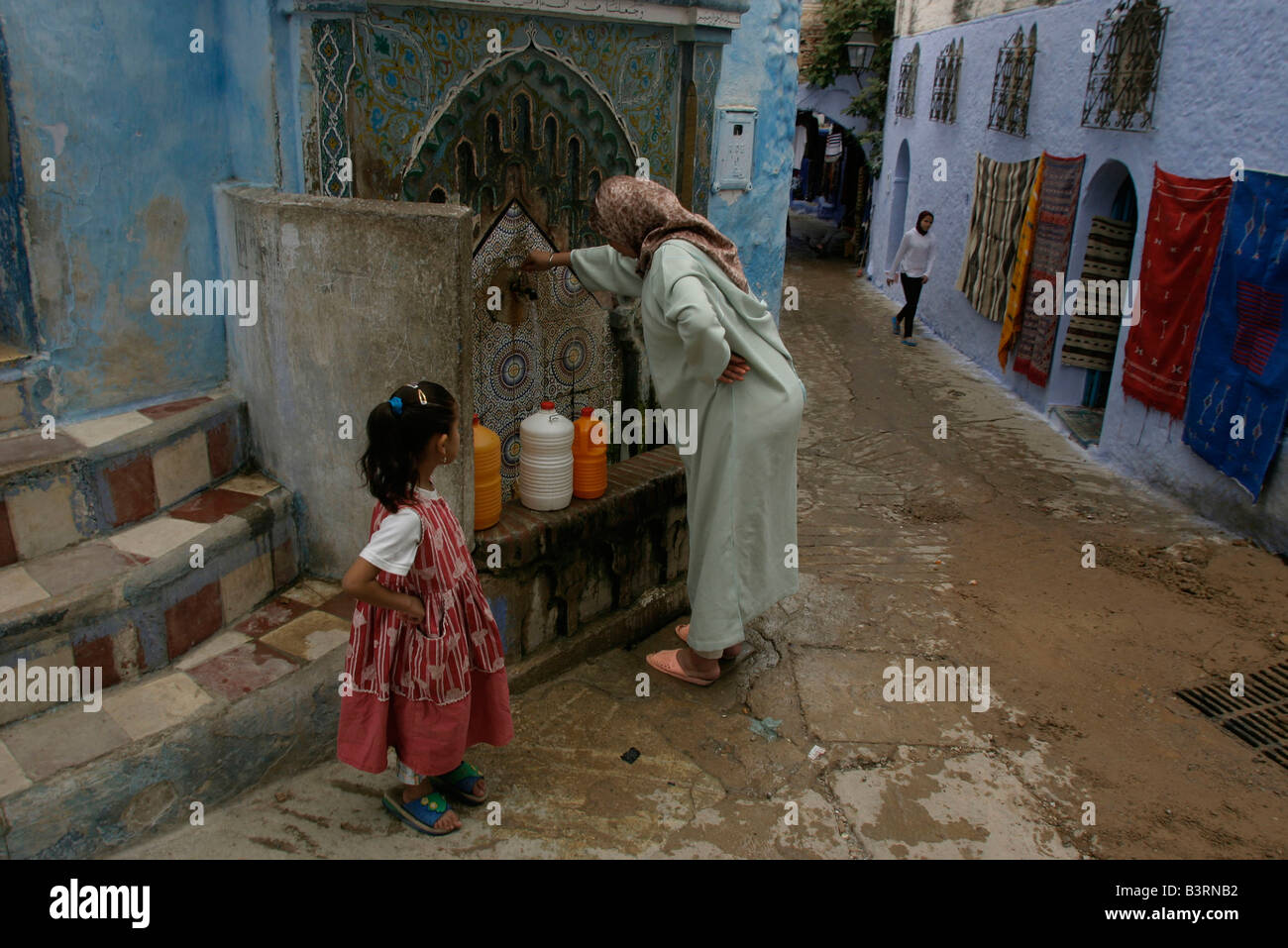 Frau füllt Wasser-Container aus Wasserhahn in der Straße von Chefchaouen. Viele Häuser in der Altstadt sind noch ohne fließendes Wasser Stockfoto