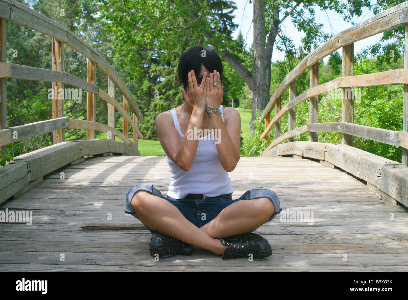Horizontale kaukasischen Brünette sitzt im Schneidersitz auf einer Brücke und bedeckte ihr Gesicht mit den Händen Stockfoto