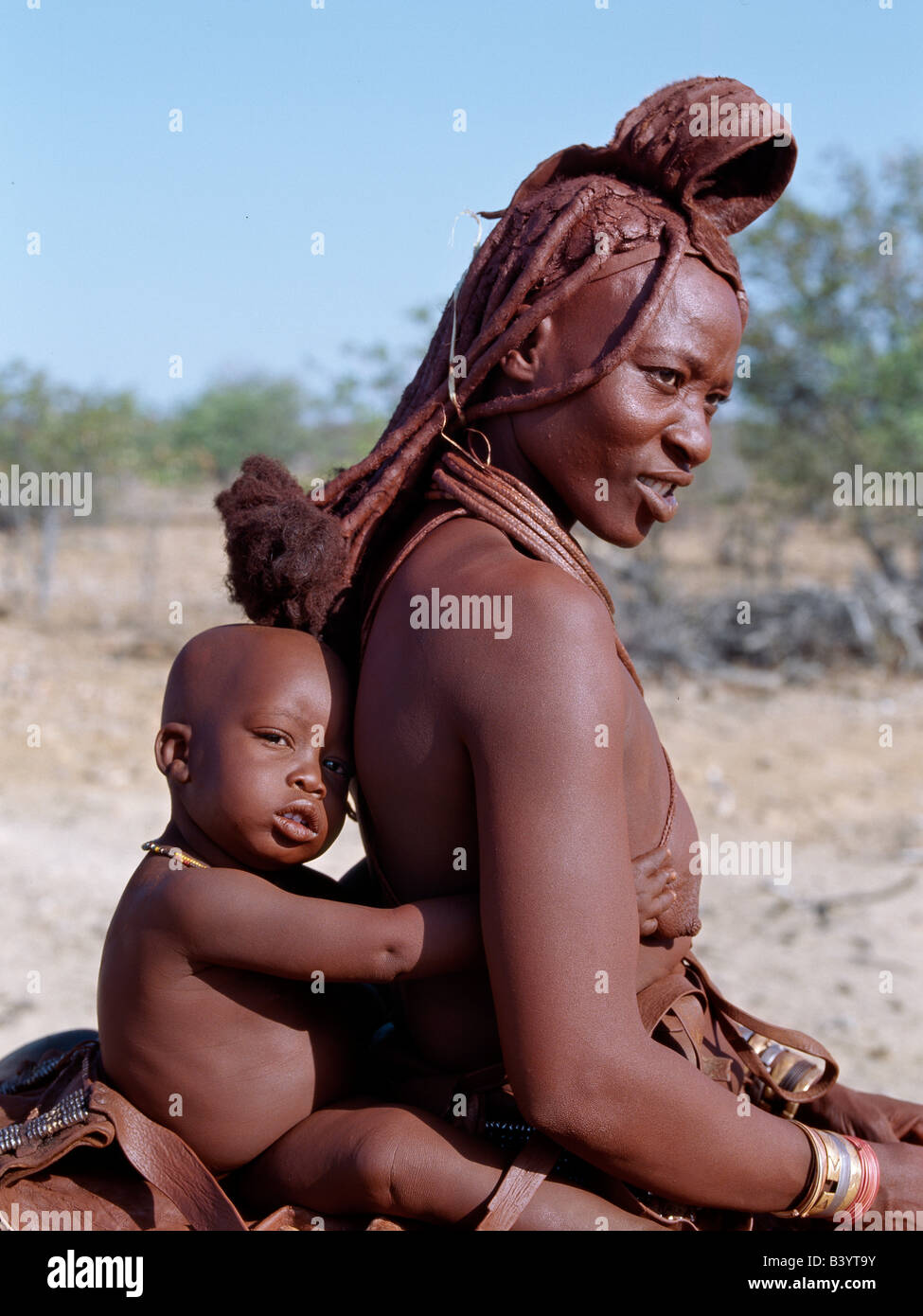Namibia, Kaokoland, Opuwo. Himba Mutter und Kind reiten nach Hause auf einem Esel. Ihre Körper glänzen aus einer Mischung von rotem Ocker, Butterfett und Kräutern. Lange Haare der Frau ist in der traditionellen Himba Weise gestaltet und wird gekrönt mit einem Kopfschmuck aus Lammfell, genannt Erembe. Die Himba sind Bantu Herero sprechende Nomaden, die in der rauen, trockenen aber krass schöne Landschaft entlegenen Nordwesten Namibias Leben. Stockfoto