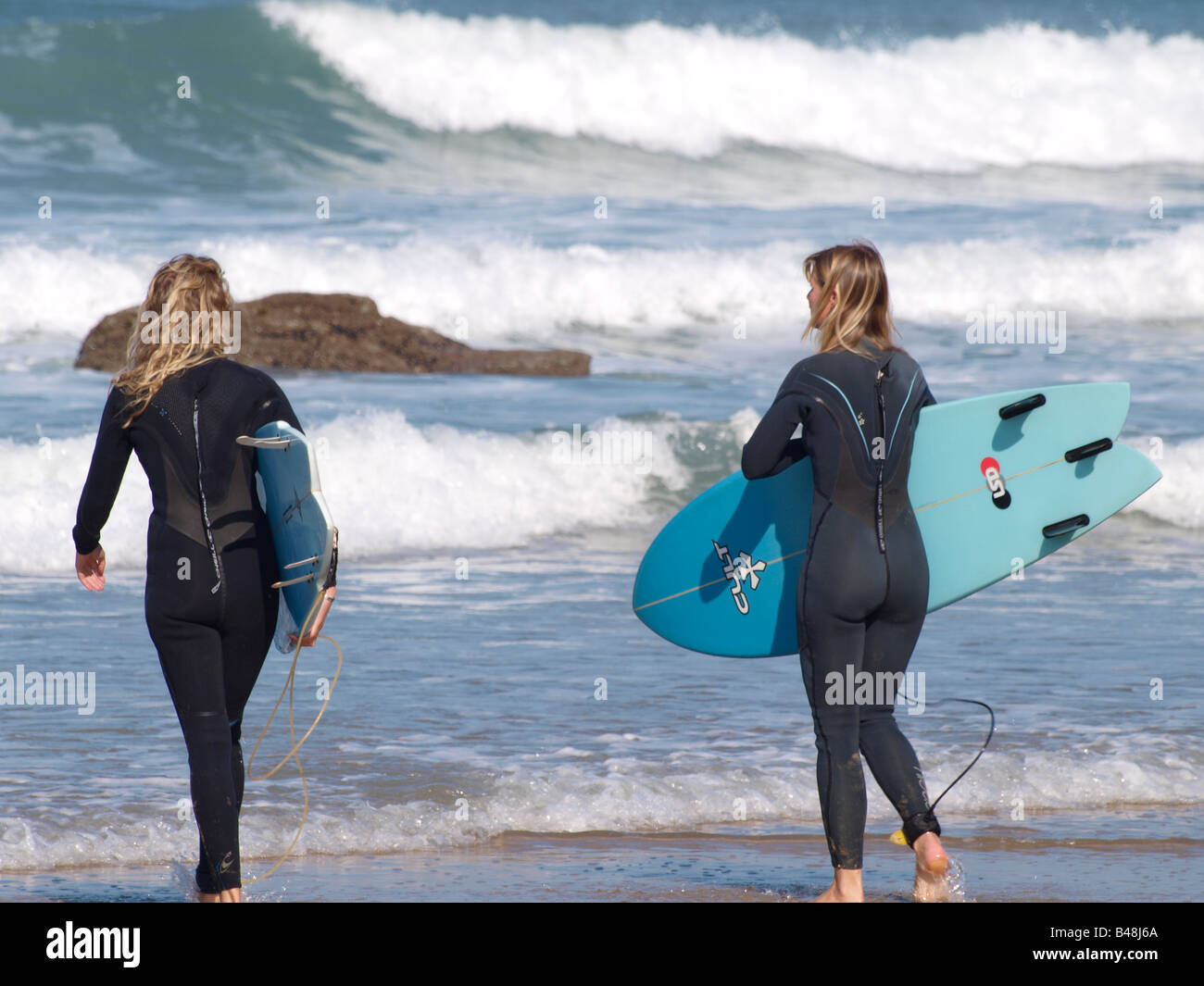 Zwei blonde weibliche Surfer ins Meer, trägt Anzüge und Surfbretter hält. Watergate Bay, Newquay, Cornwall Stockfoto
