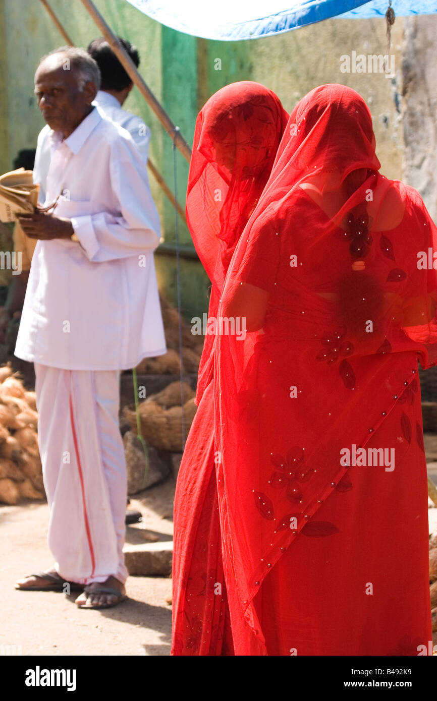 Frauen in roten Saris und Schleier besuchen einen Straßenmarkt in Mysore, Indien. Im Hintergrund steht ein Mann in einem weißen Anzug. Stockfoto