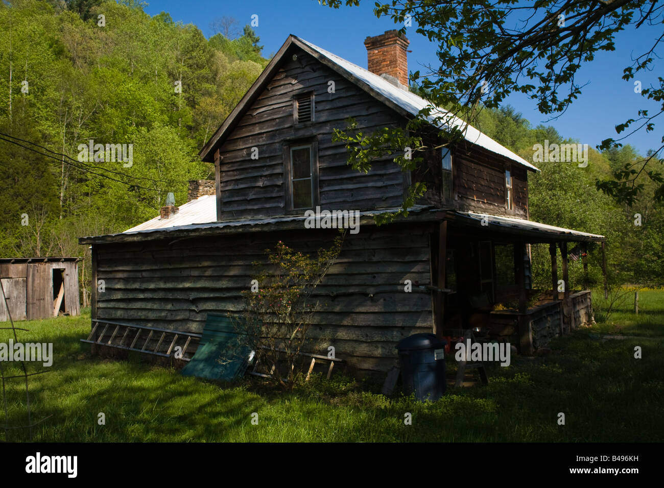 Schatten von Bäumen nähern sich ein alten hölzernen Bauernhaus mit einem Blechdach. Ländliche Umgebung, am Abend mit einem Wald. Stockfoto